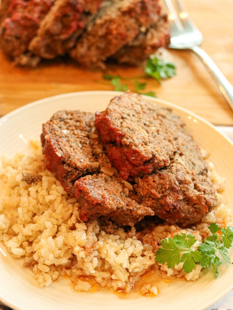 Two slices of eggless meatloaf on a plate with rice and a sprig of parsley with more meatloaf in the background.