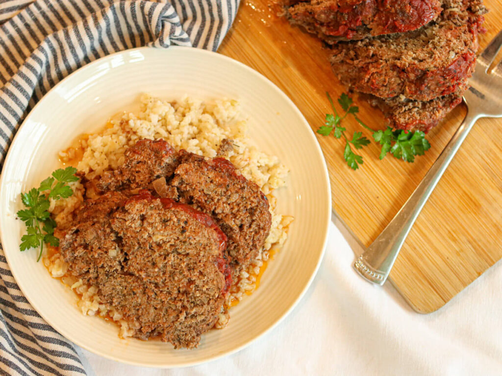 Two slices of meatloaf on a plate with rice on a white tablecloth with a blue and white napkin.