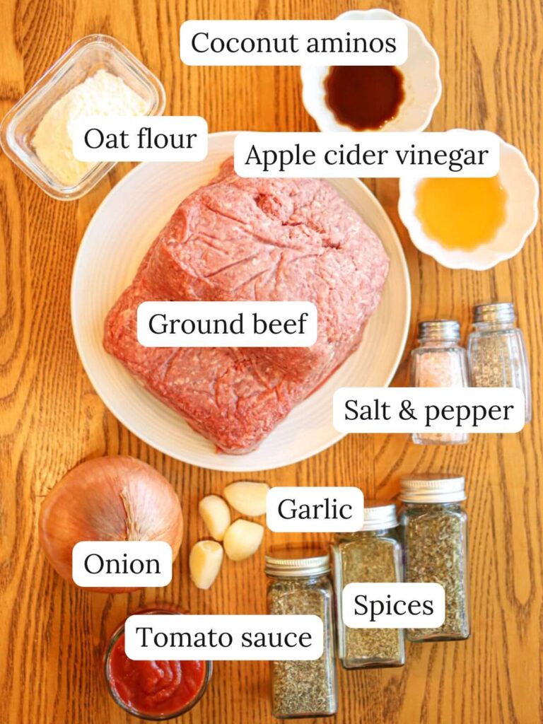 Labeled ingredients on a wooden table including ground beef, onion, garlic, spices, tomato sauce, coconut aminos, and oat flour.
