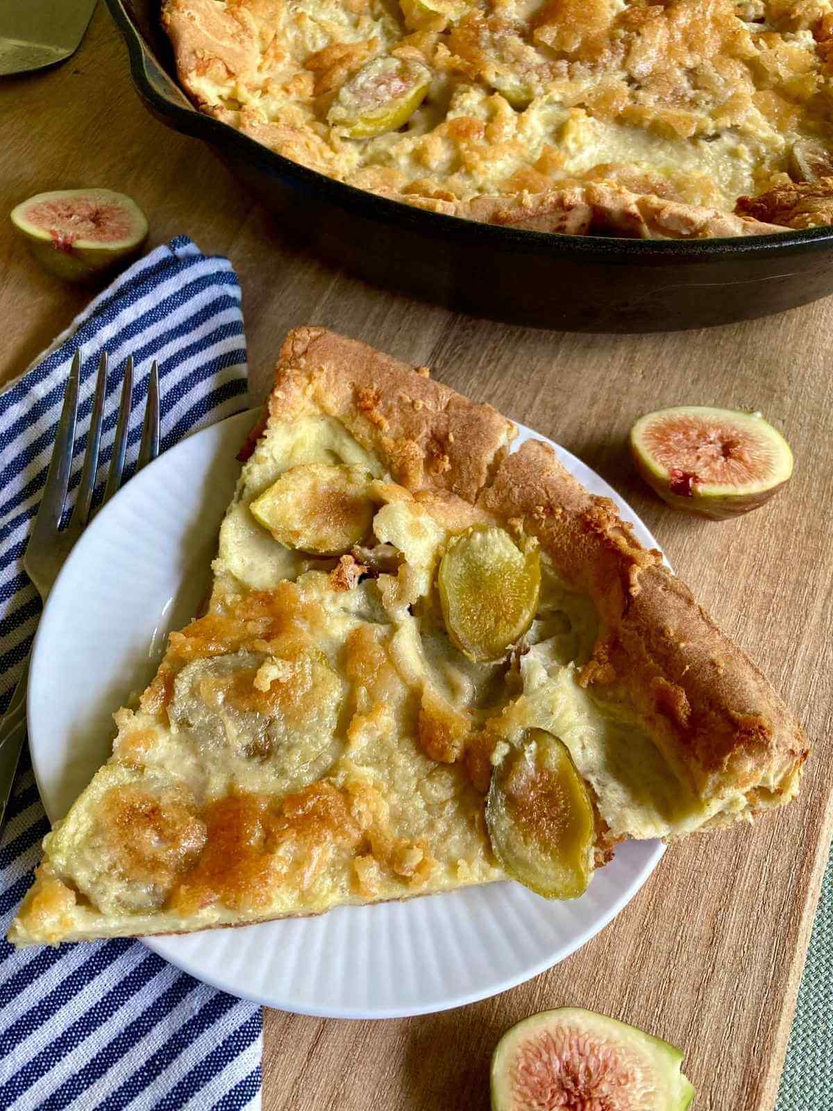 Slice of fig Dutch baby on a plate with a skillet with the rest of the dutch baby in the background.