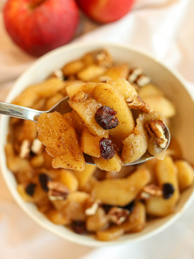 A spoon scooping fried apples topped with cranberries and pecans out of a bowl.