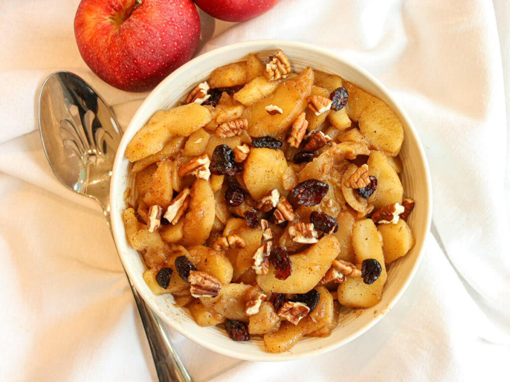 A bowl with fried apples on a white tablecloth beside fresh apples and a serving spoon.