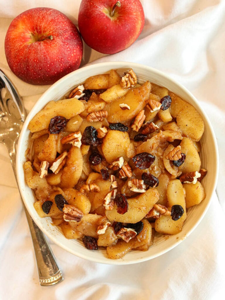 A bowl full of healthy fried apples on a white tablecloth beside fresh apples and a serving spoon.