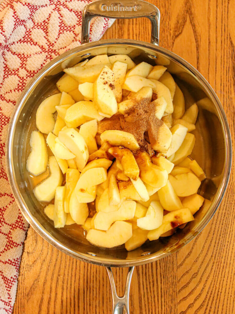 Apple slices, spices, and other ingredients in a saute pan on a wooden surface.