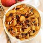 Healthy fried apples topped with pecans and cranberries in a white serving bowl on a tablecloth beside fresh apples and a serving spoon.