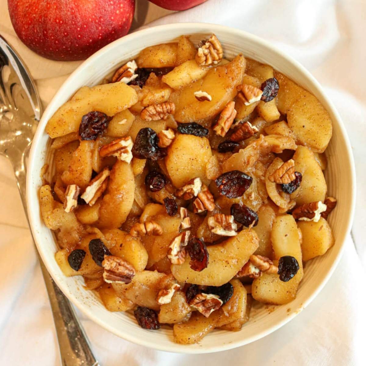 Healthy fried apples topped with pecans and cranberries in a white serving bowl on a tablecloth beside fresh apples and a serving spoon.