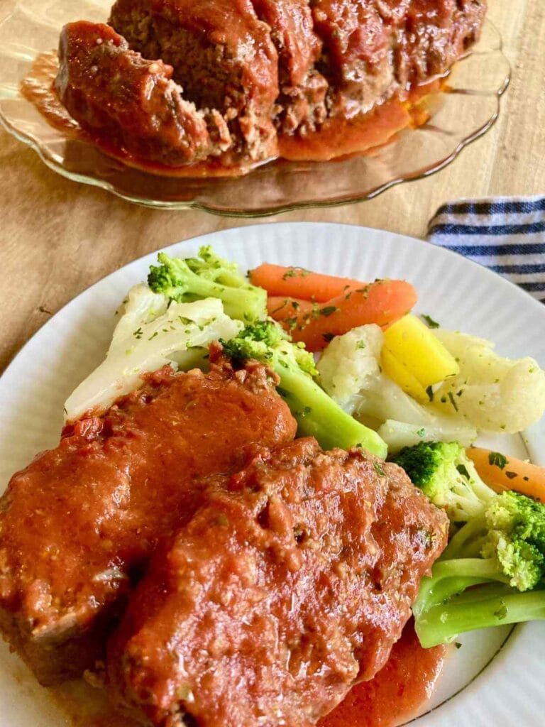 Sliced meatloaf on a serving platter and two slices on a plate with steamed vegetables.