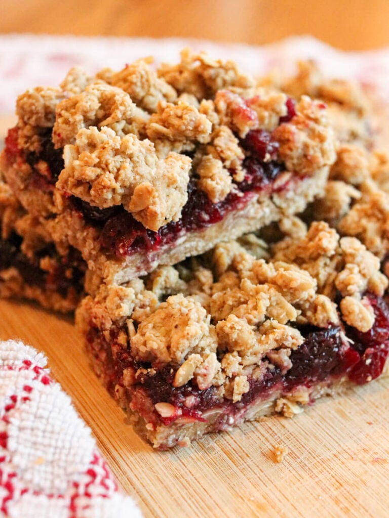 Gluten-free cranberry bars with cranberry sauce stacked on a wooden cutting board beside a red and white kitchen cloth.