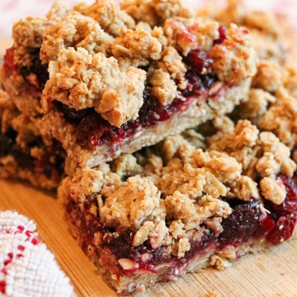 Closeup of gluten-free cranberry crips bars with cranberry sauce stacked on a wooden cutting board beside a red and white kitchen cloth.