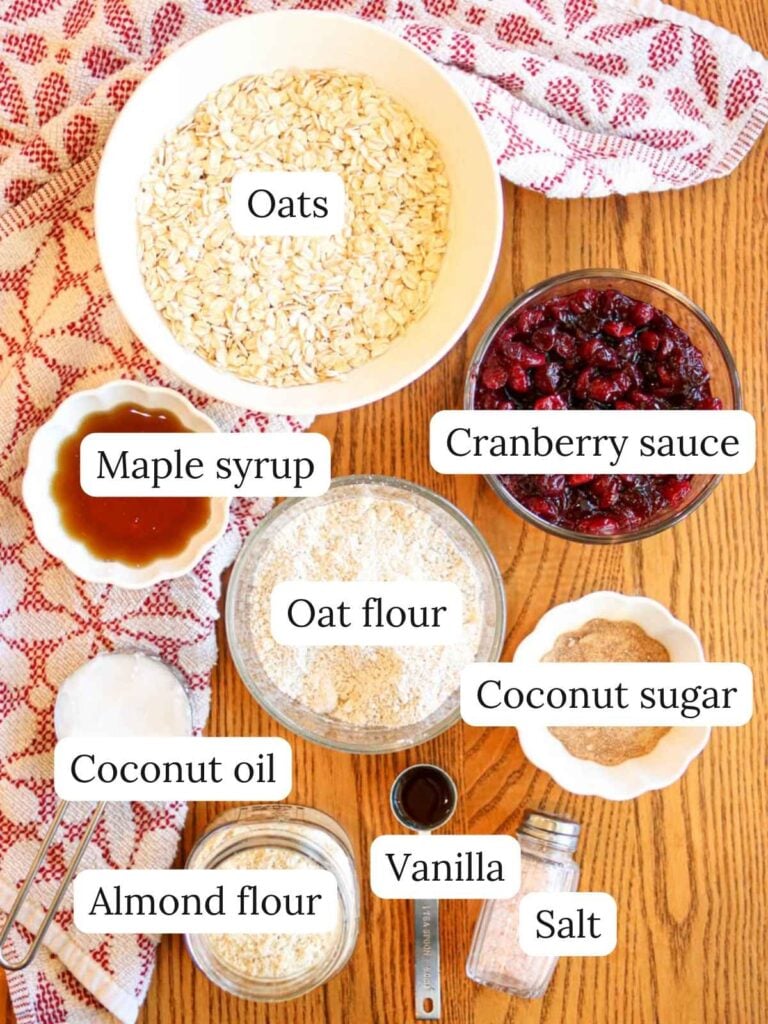 Labeled ingredients on a table with a red and white kitchen cloth including oats, cranberry sauce, maple syrup, oat flour, coconut sugar, coconut oil, almond flour, vanilla, and salt.
