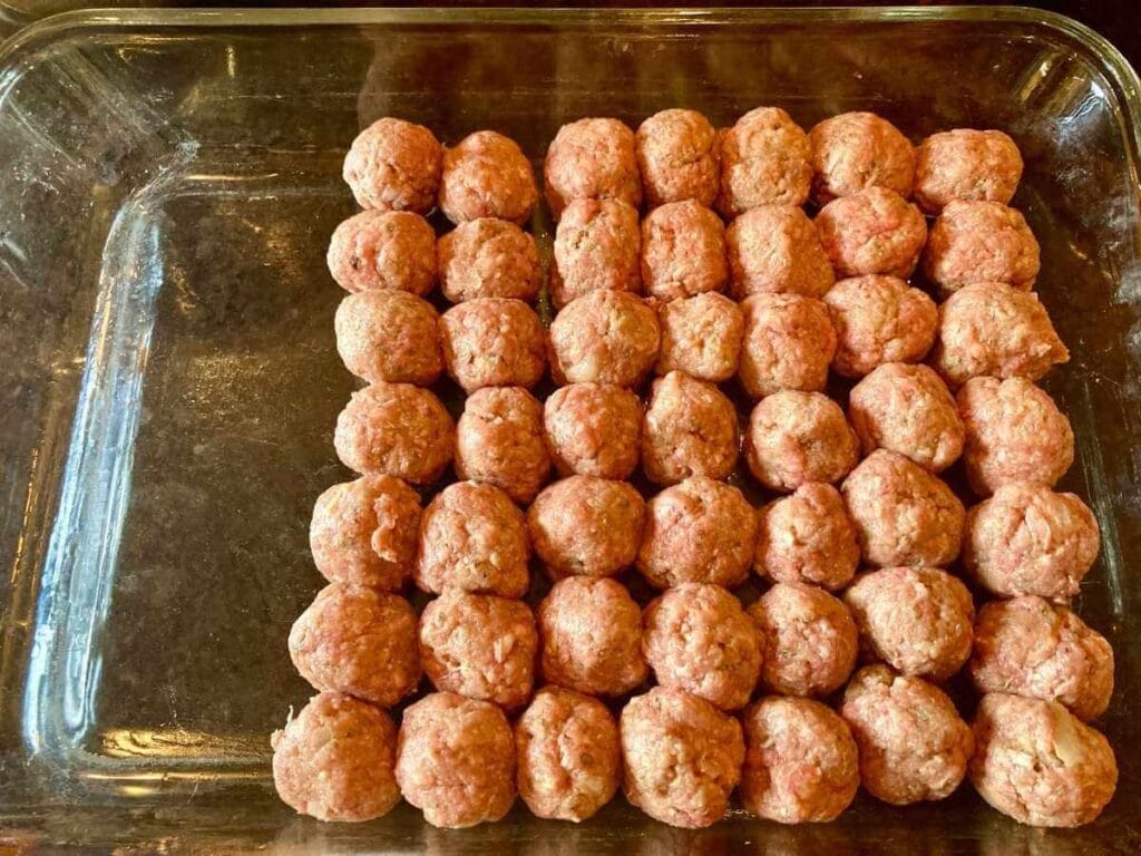 Glass baking dish half filled with uncooked meatballs.