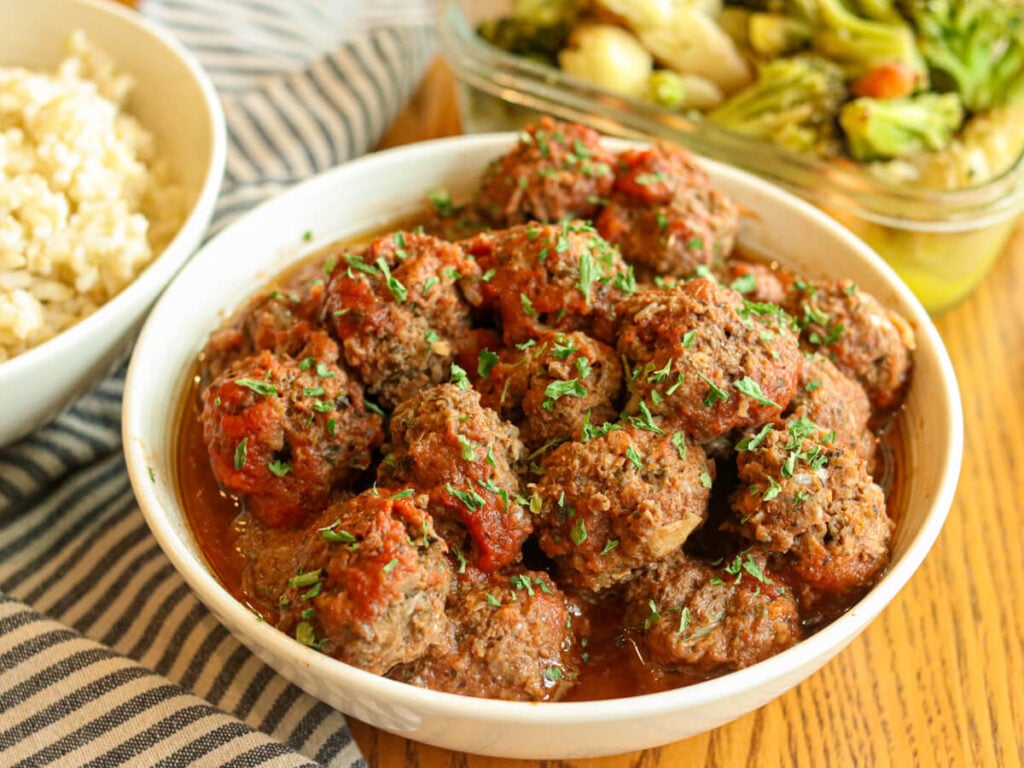 Breadcrumb-free meatballs in a serving bowl on a table with bowls of rice and vegetables in the background.