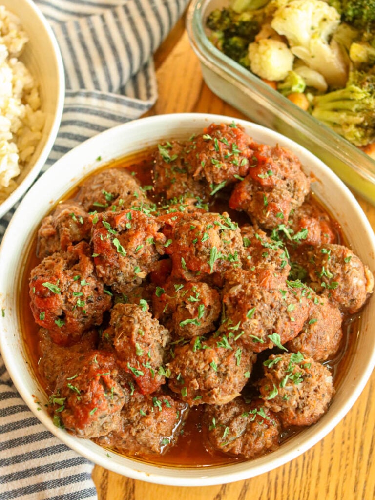 Meatballs in a white bowl on a table with bowls of rice and vegetables in the background.