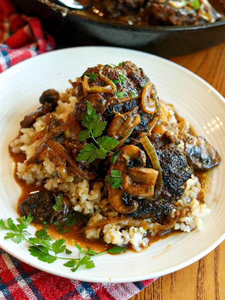 Two Salisbury steak patties with mushroom and onion gravy served over rice on a white plate garnished with fresh parsley.