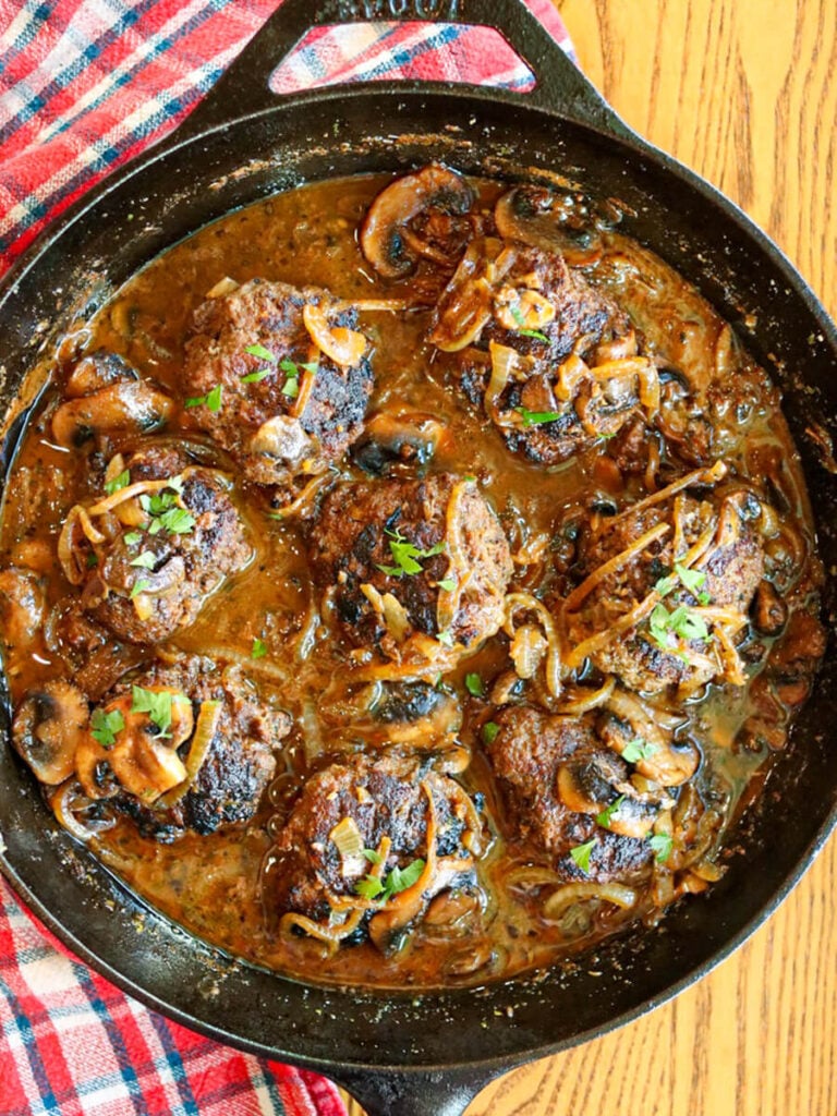 Gluten-free Salisbury steak patties in a cast iron skillet on a wooden surface beside a checkered cloth.