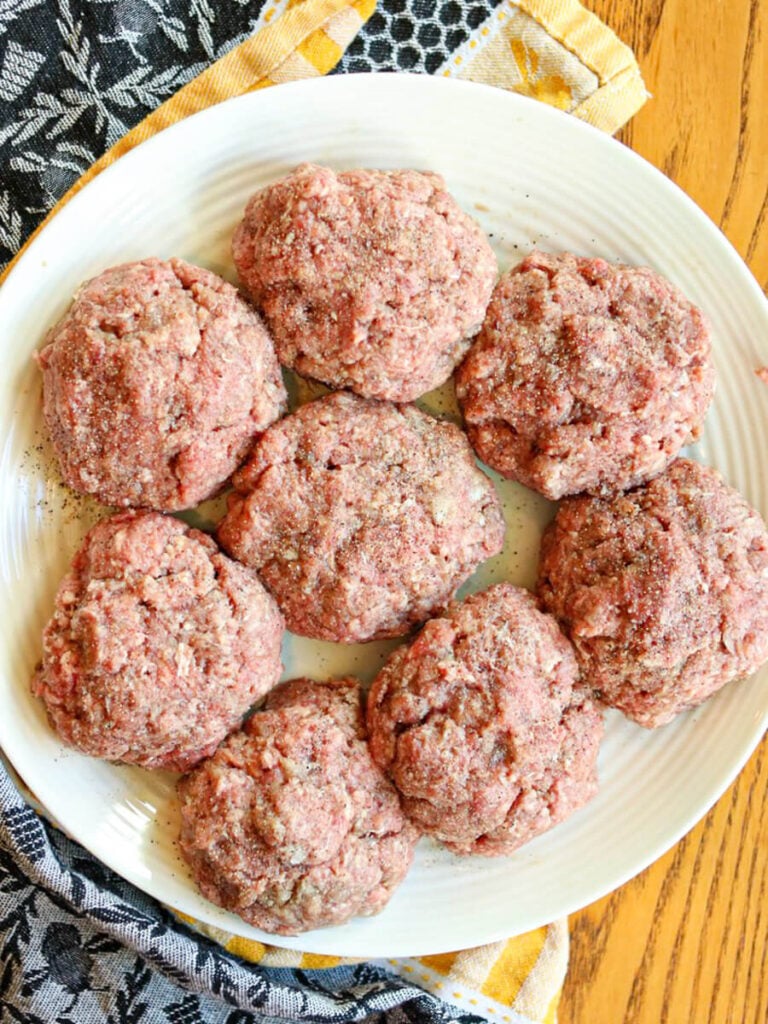 Raw beef patties on a white plate on top of a kitchen towel.