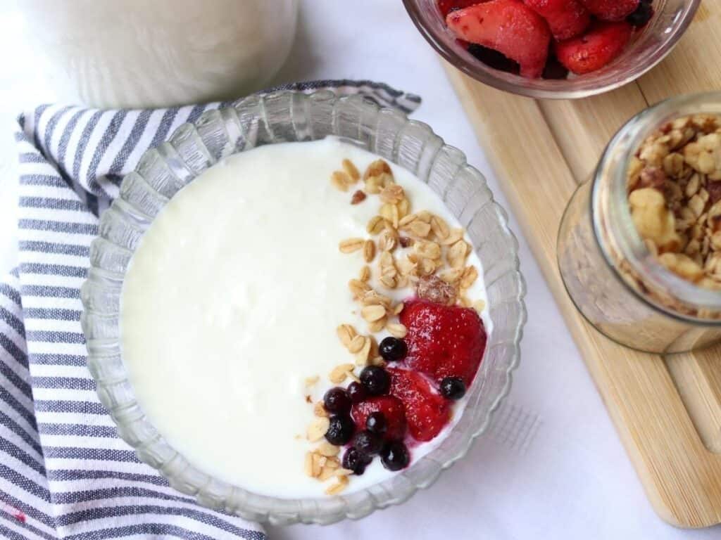 A glass bowl of homemade yogurt topped with granola, strawberries, and blueberries on a striped napkin. Nearby are jars of granola, fresh berries, and yogurt.