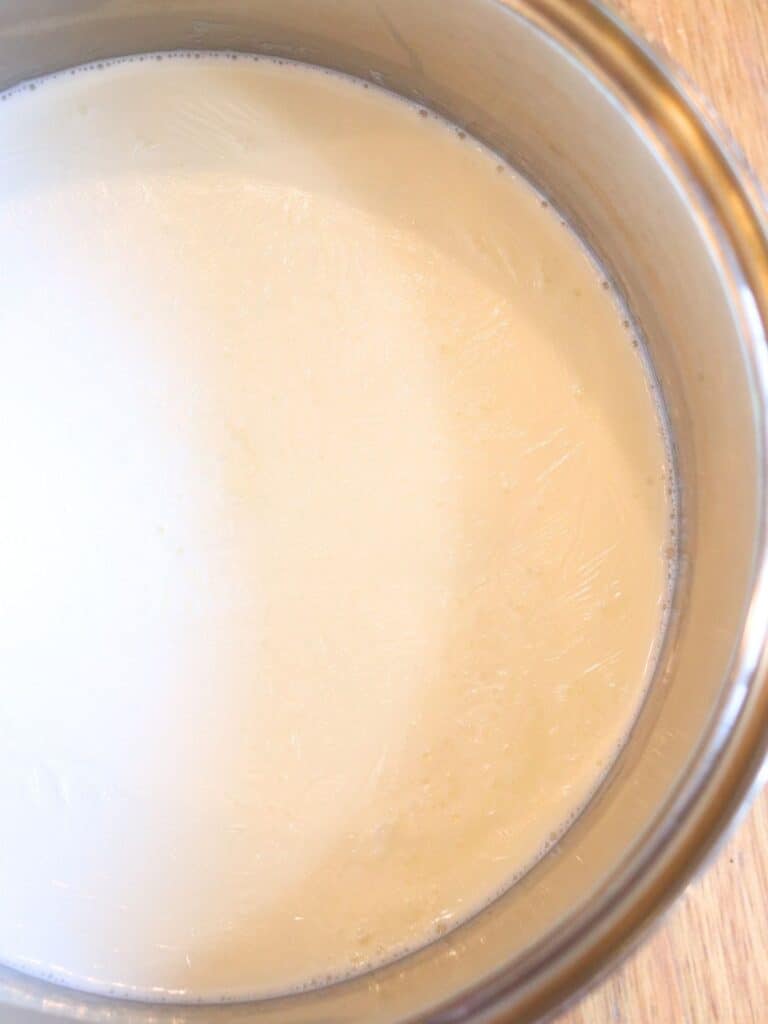 A close-up of a shiny stainless steel pot filled with heated milk, slightly frothy along the edges, resting on a light wooden surface.
