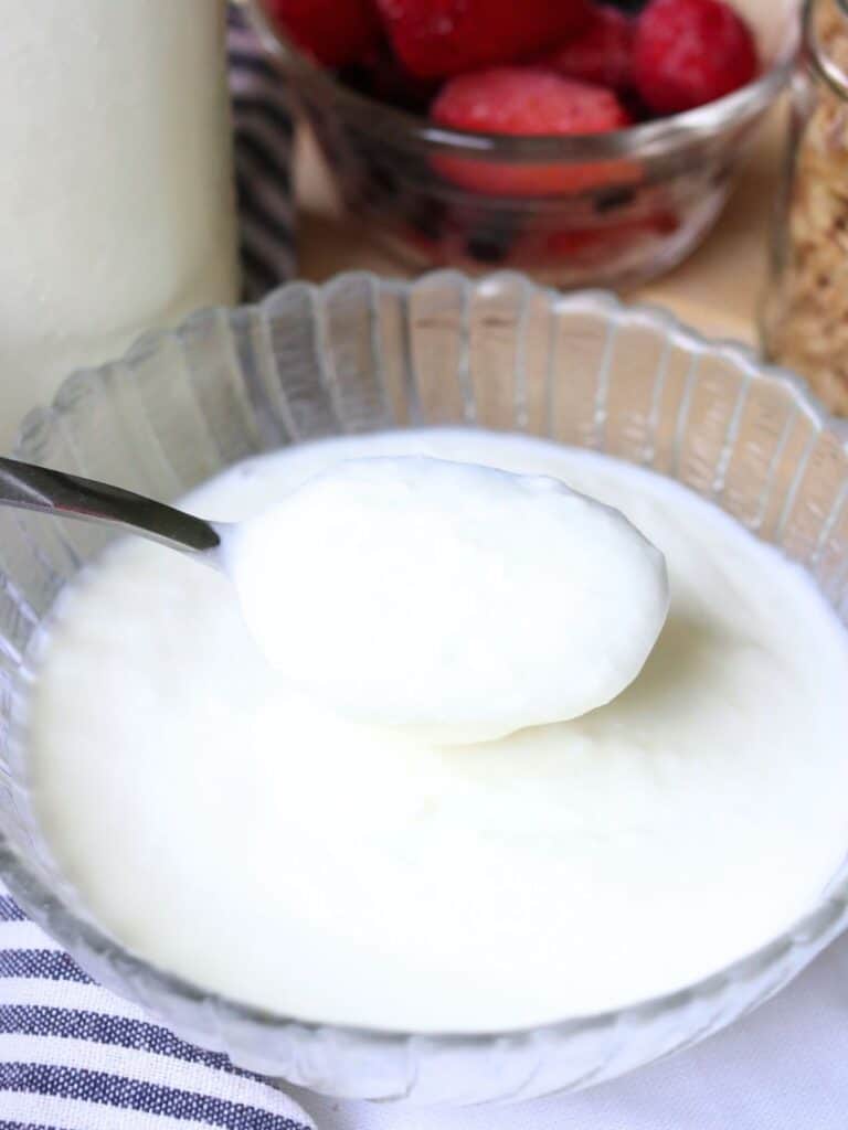 A spoonful of creamy, white homemade yogurt is held above a glass bowl, with fresh berries in a bowl behind it. The scene is bright and inviting.