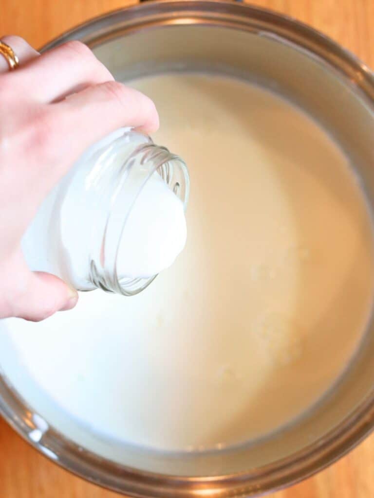 Pouring yogurt starter from a glass jar into a stainless steel pot of warm milk.