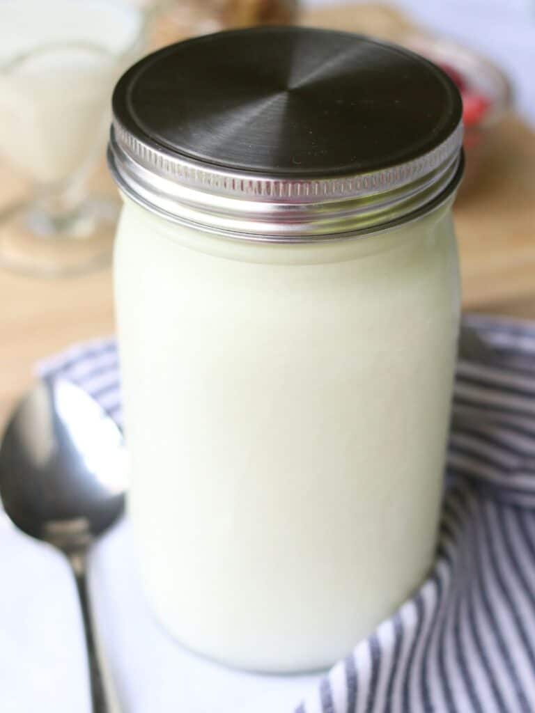 Glass jar full of fresh and creamy homemade yogurt with a spoon and striped cloth napkin beside it.