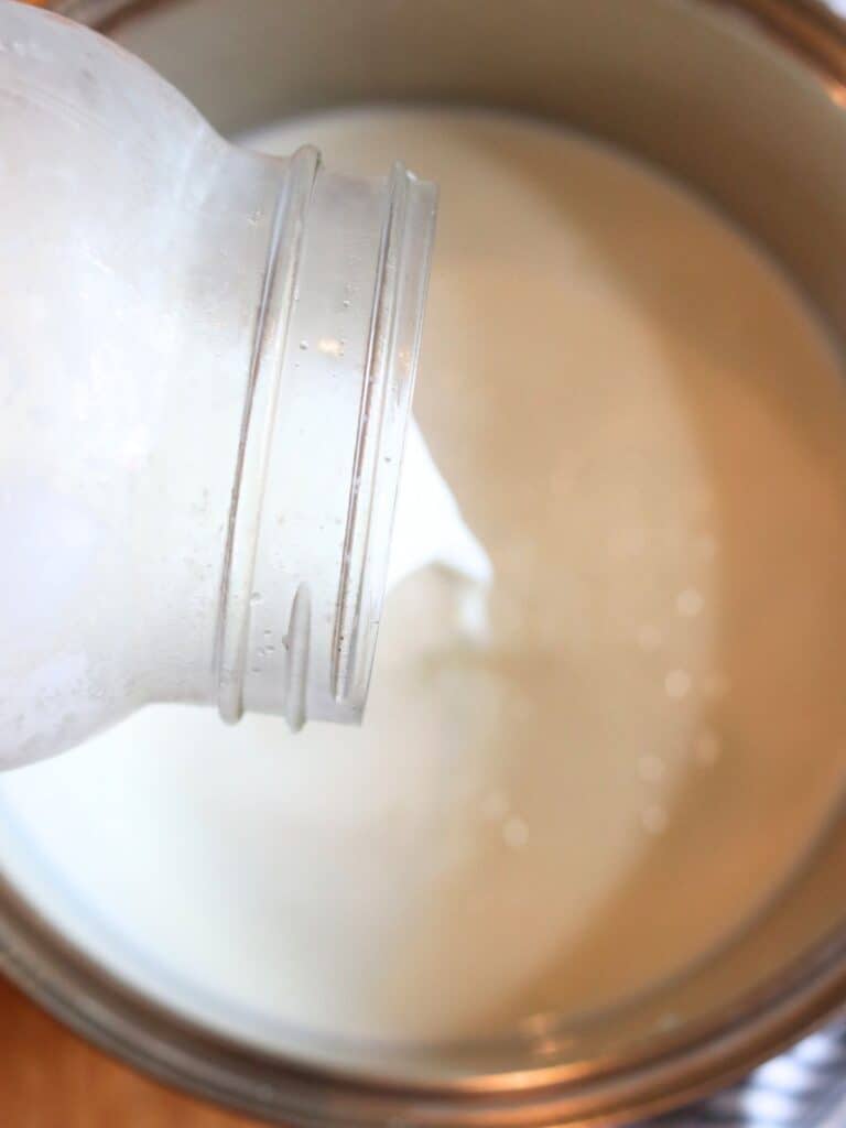 The first step of making yogurt - pouring milk from a glass jar into a metal bowl, with creamy texture and soft lighting. The image conveys freshness and simplicity.