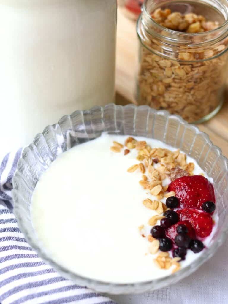 A glass bowl of yogurt made from raw milk topped with granola, strawberries, and blueberries on a striped napkin. Nearby is a jar of granola.