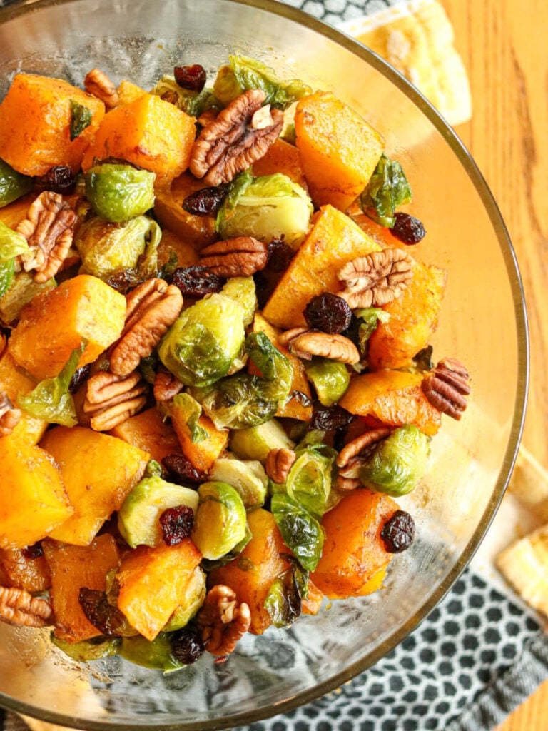 View of half of a glass bowl with roasted Brussels sprouts and butternut squash topped with cranberries and pecans on a wooden table with a kitchen cloth.