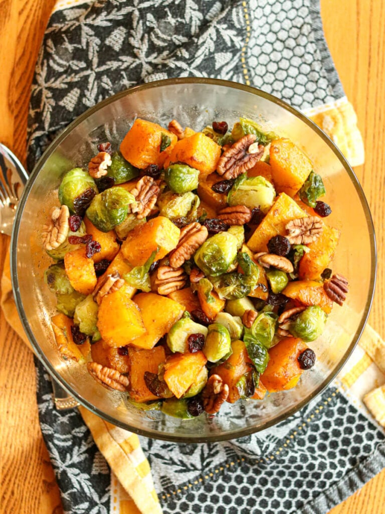 Glass serving bowl filled with roasted Brussels sprouts and butternut squash topped with cranberries and pecans on a wooden table with a kitchen cloth.