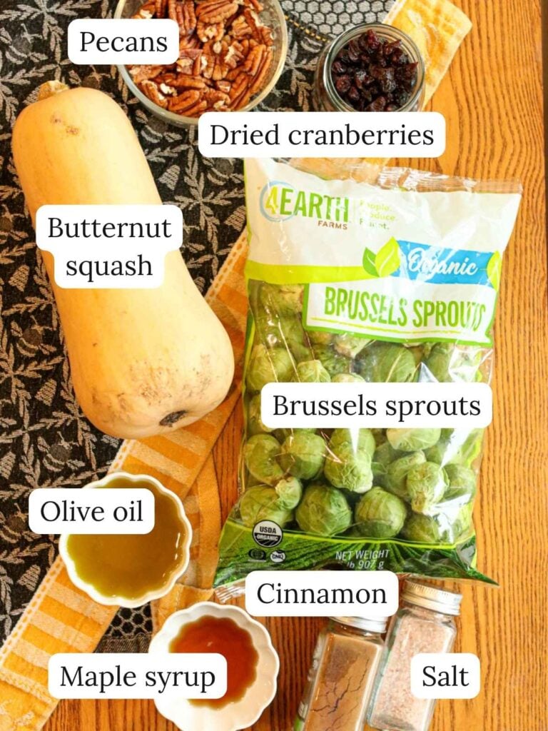 Labeled ingredients on a wood table with a navy and yellow towel including pecans, butternut squash, dried cranberries, Brussels sprouts, olive oil, maple syrup, cinnamon, and salt.