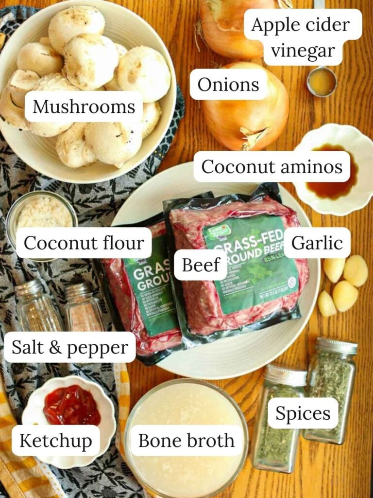 Labeled ingredients on a wood surface with a kitchen cloth including mushrooms, onions, apple cider vinegar, coconut aminos, coconut flour, beef, garlic, salt and pepper, ketchup, bone broth, and spices.