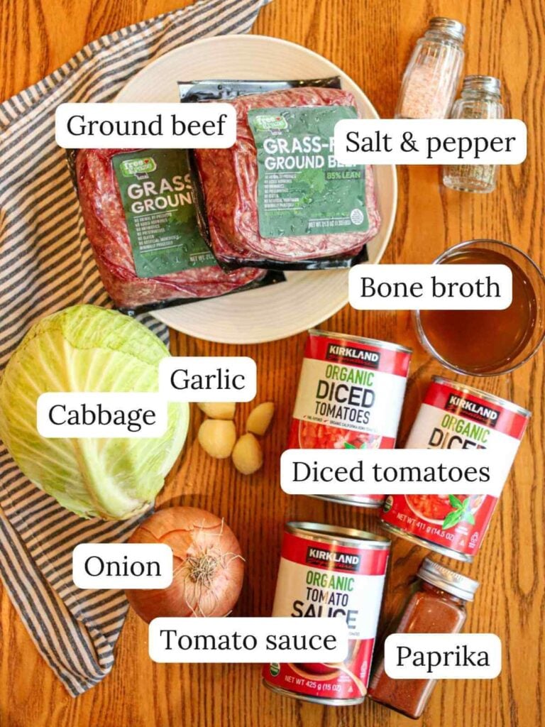 Labeled ingredients on a wooden table including ground beef, salt and pepper, cabbage, garlic, diced tomatoes, onion, tomato sauce, paprika, and bone broth.