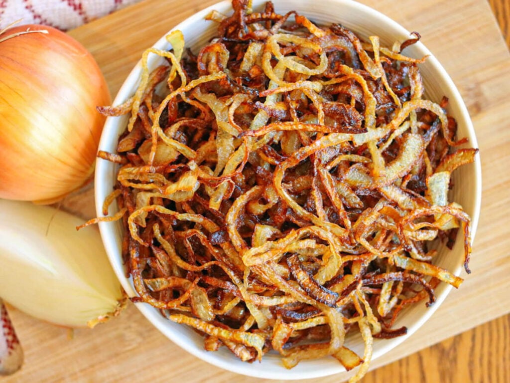 A bowl of gluten-free fried onions in a white bowl on a wooden cutting board with fresh onions in the background.