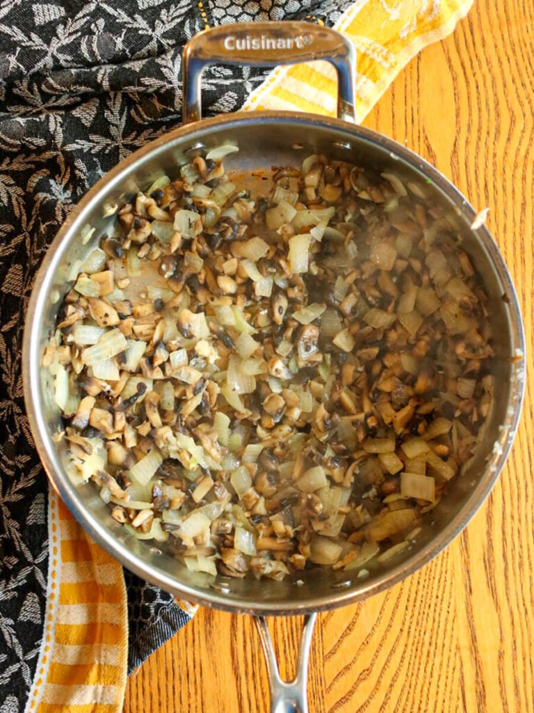 Onions, mushrooms, and garlic sautéing in a stainless steel pan on a wooden background.