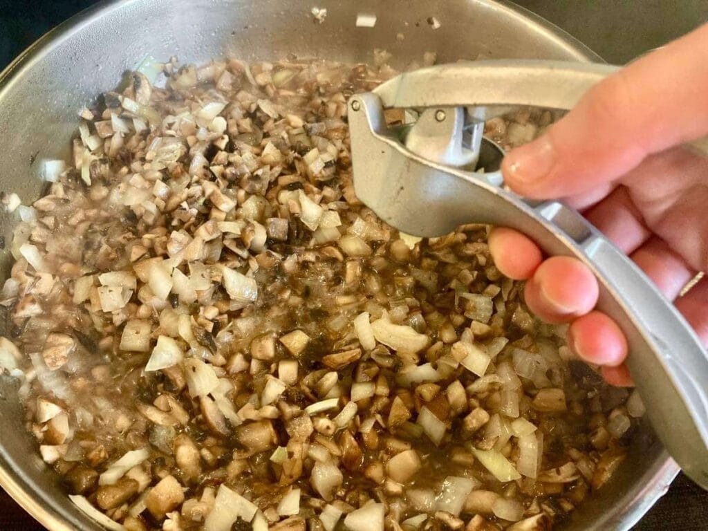 Crushing garlic into skillet with mushrooms and onions