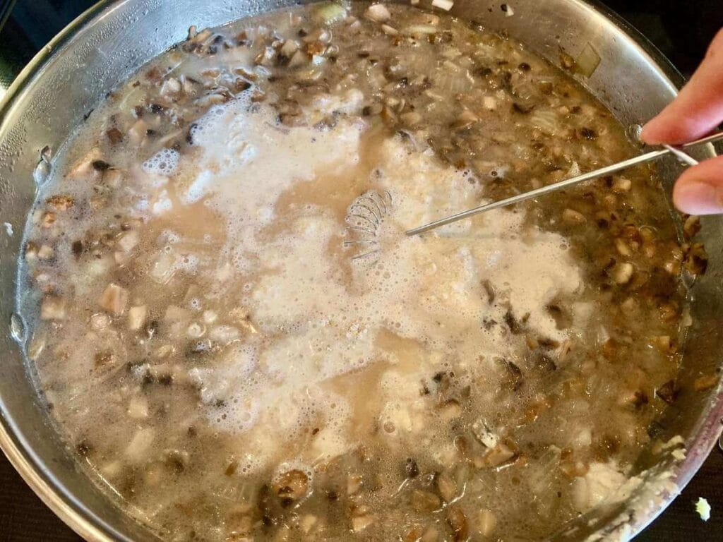 Whisking broth, cream, and oat flour into mushrooms and onions in skillet