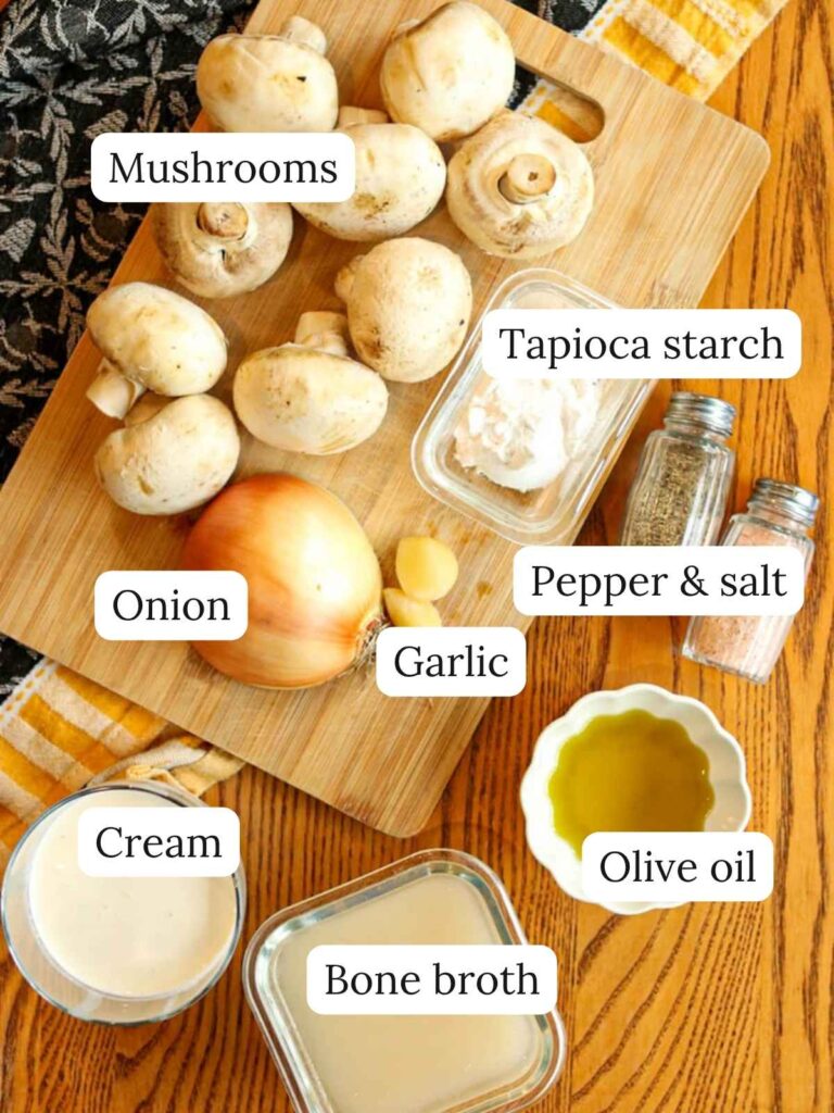 Labeled soup ingredients on a wooden table and cutting board including mushrooms, onion, tapioca starch, garlic, salt, pepper, cream, bone broth, and olive oil.