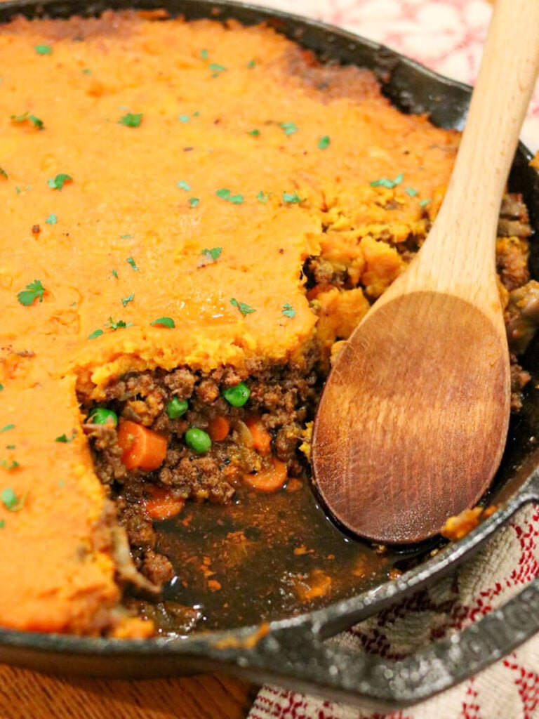A wooden spoon in a cast iron skillet with sweet potato cottage pie with a piece taken out.
