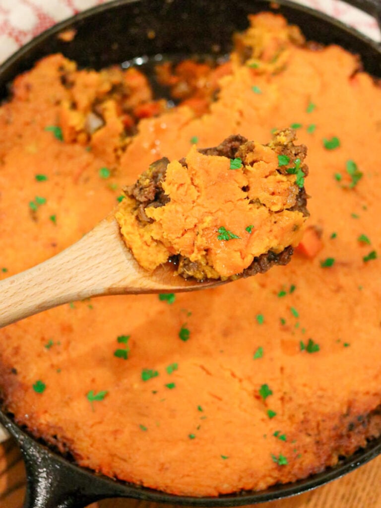 A wooden spoon holding a scoop of sweet potato cottage pie above the dish.
