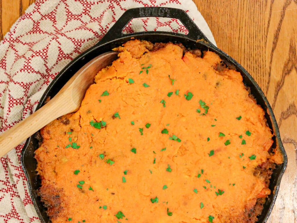 A cast iron skillet filled with sweet potato cottage pie with a wooden spoon in it.