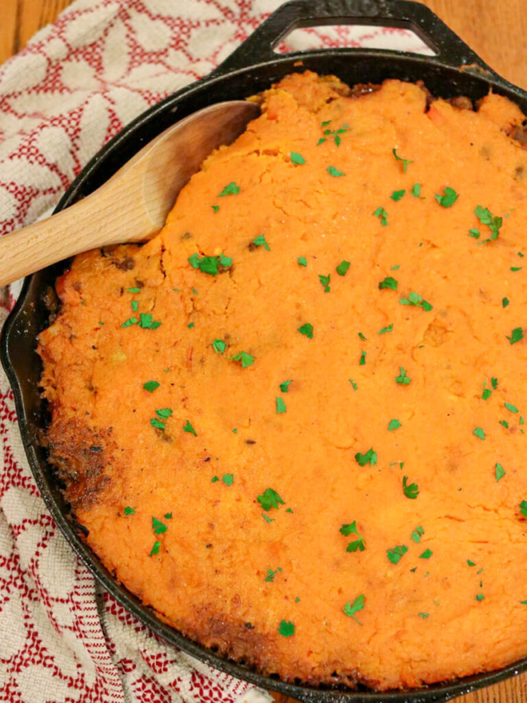 A cast iron skillet filled with sweet potato cottage pie with a wooden spoon in it surrounded by a kitchen towel.