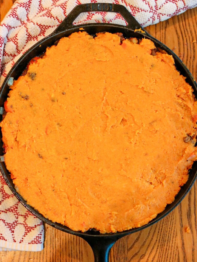 Mashed sweet potatoes spread over meat and vegetable mixture in a skillet on a wooden table.