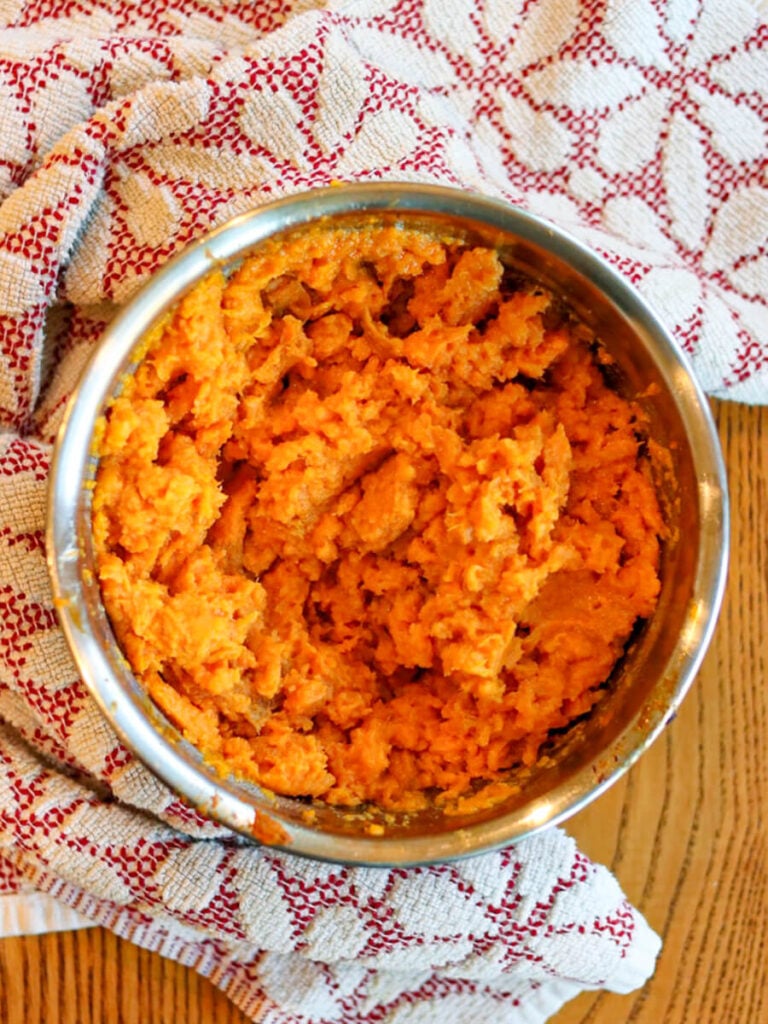 Mashed sweet potatoes in a metal mixing bowl surrounded by a kitchen towel.