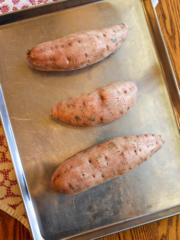 Three sweet potatoes on a stainless steel baking sheet ready to bake.