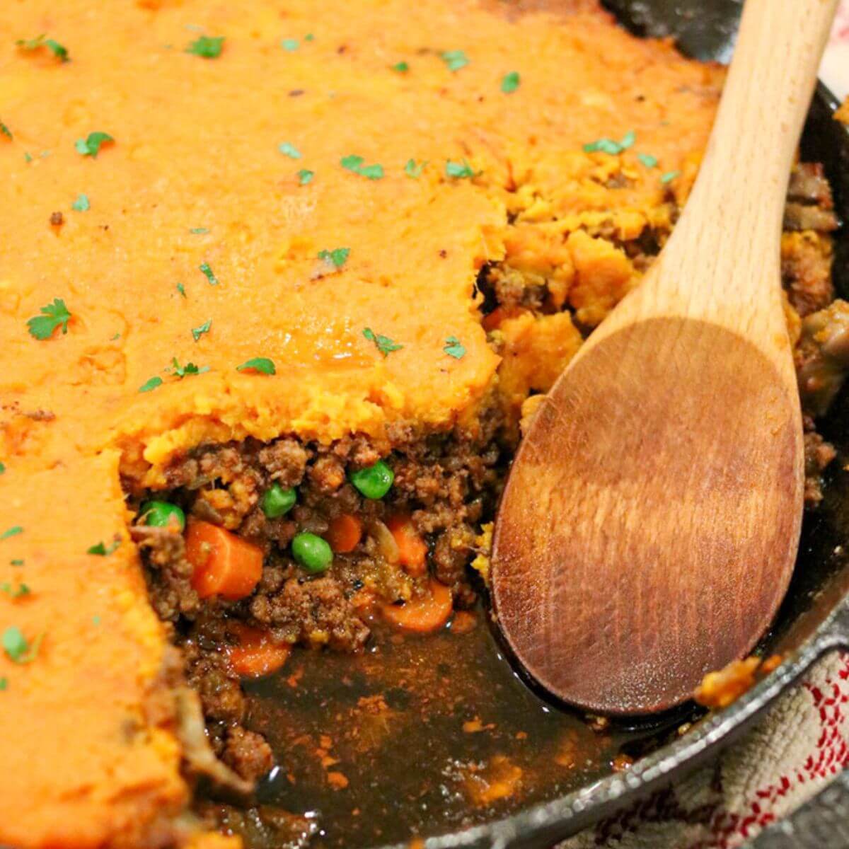 Closeup view of a wooden spoon in a cast iron skillet with sweet potato cottage pie with a piece taken out.