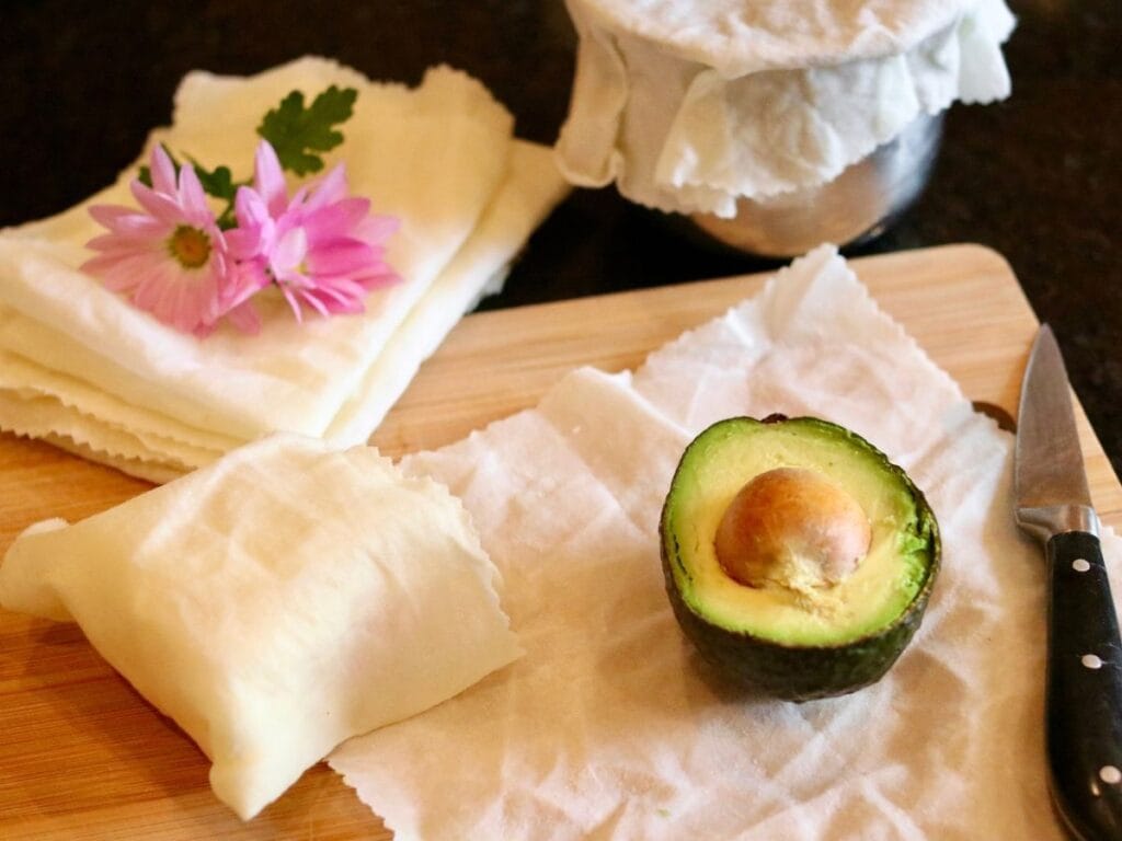 Cheese wrapped in beeswax food wrap with half of an avocado on a piece of beeswax wrap with a covered bowl and stack of food wraps.