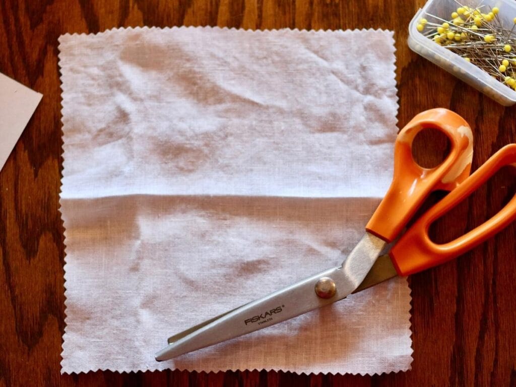 Square piece of white linen fabric with pinking shears and pins.
