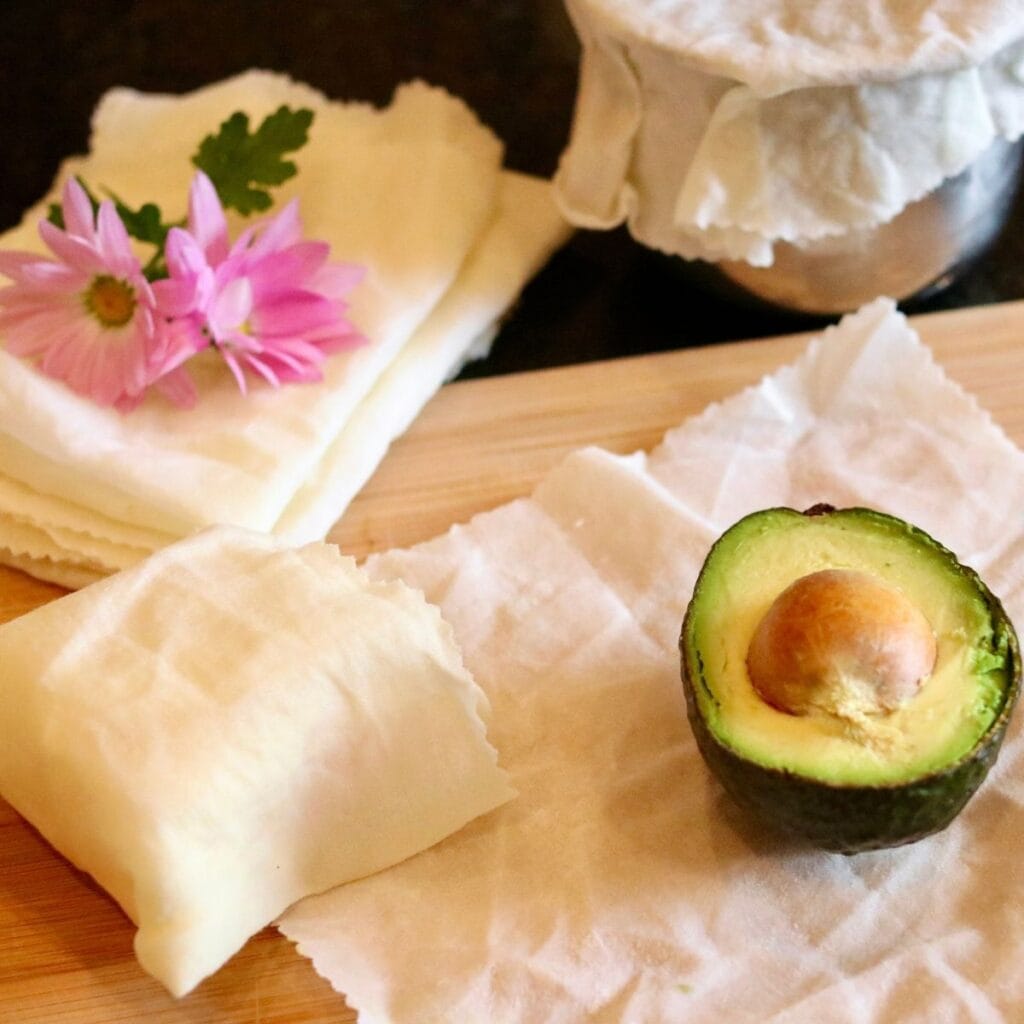 Closeup of cheese wrapped in beeswax food wrap with half of an avocado on a piece of beeswax wrap with a covered bowl and stack of food wraps.