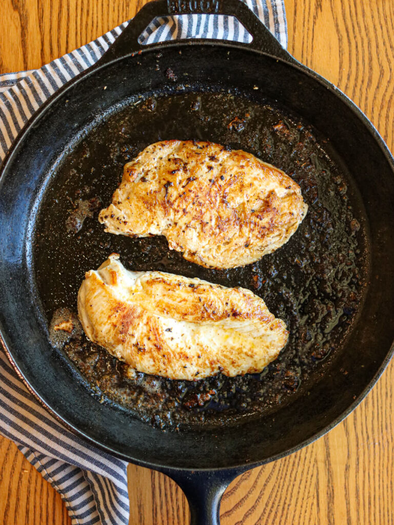 Two chicken breasts searing in a cast iron skillet.
