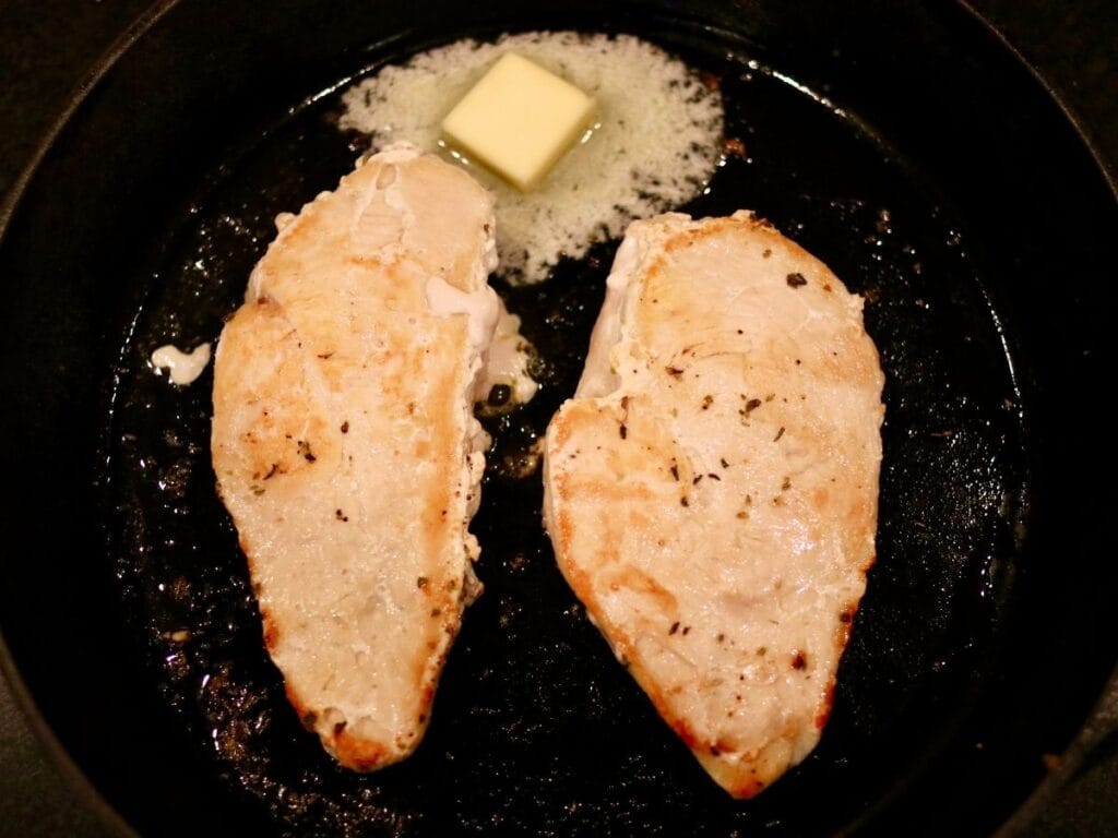 2 chicken breasts cooking with butter in a cast iron skillet